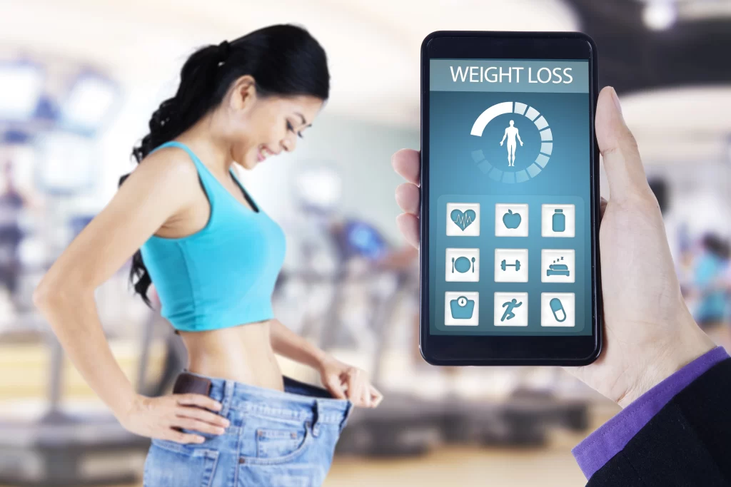 A conceptual image contrasting physical fitness results with digital tracking. In the foreground, a hand holds a smartphone displaying a "Weight Loss" app interface with a progress meter and fitness icons. In the blurred background, a smiling woman, who has clearly lost weight, holds up the waistband of her now-oversized old jeans while standing in a gym.