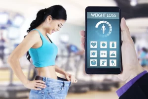 A conceptual image contrasting physical fitness results with digital tracking. In the foreground, a hand holds a smartphone displaying a "Weight Loss" app interface with a progress meter and fitness icons. In the blurred background, a smiling woman, who has clearly lost weight, holds up the waistband of her now-oversized old jeans while standing in a gym.