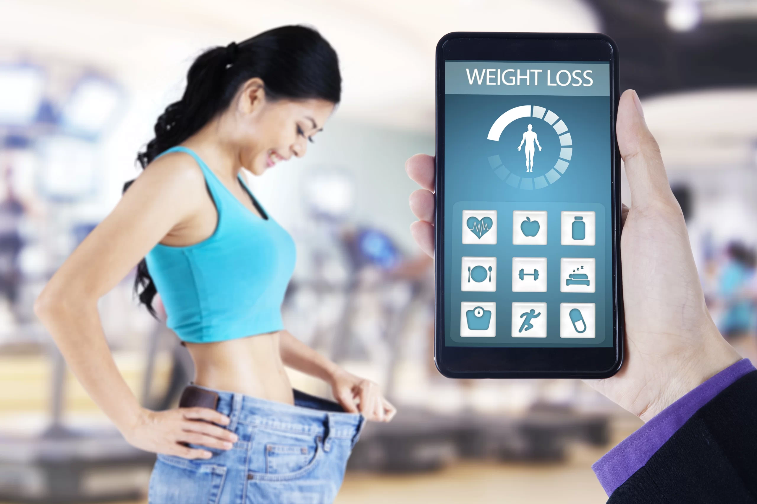 A conceptual image contrasting physical fitness results with digital tracking. In the foreground, a hand holds a smartphone displaying a "Weight Loss" app interface with a progress meter and fitness icons. In the blurred background, a smiling woman, who has clearly lost weight, holds up the waistband of her now-oversized old jeans while standing in a gym.